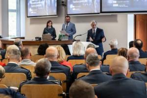 Przemawiający przy mównicy prof. Marek Walancik, audytorium, w tle dwóch prowadzących wydarzenie oraz plakat sympozjum wyświetlany na ekranach
