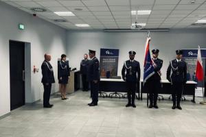 Dwóch funkcjonariuszy w niebieskim umundurowaniu stoi naprzeciwko siebie. Jeden z nich składa meldunek. Za jego plecami stoi trzech funkcjonariuszy ze sztandarem.
