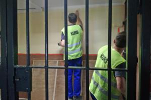 Dwóch mężczyzn w kamizelkach z napisem KURS maluje ścianę, przed nimi jest krata.
